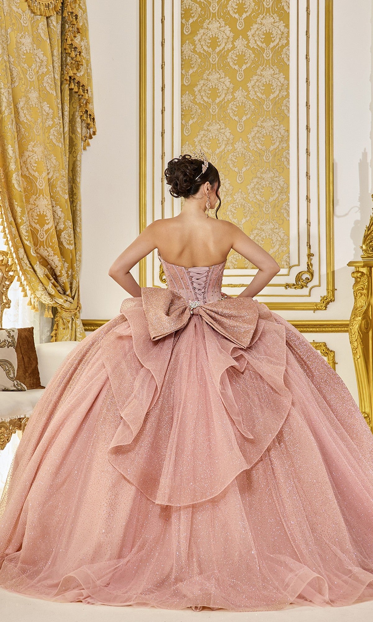 A woman stands facing away in an ornate gold and white room, wearing the Quinceanera Dress 5078 By Dancing Queen—a pink, strapless corset bodice ball gown with a large bow.